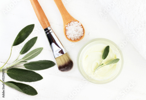  Herbal botanical facial mask with salvia. Ingredients for home spa top view: Jar of cream, green plant leaves, sea salt, white wooden table background. 
