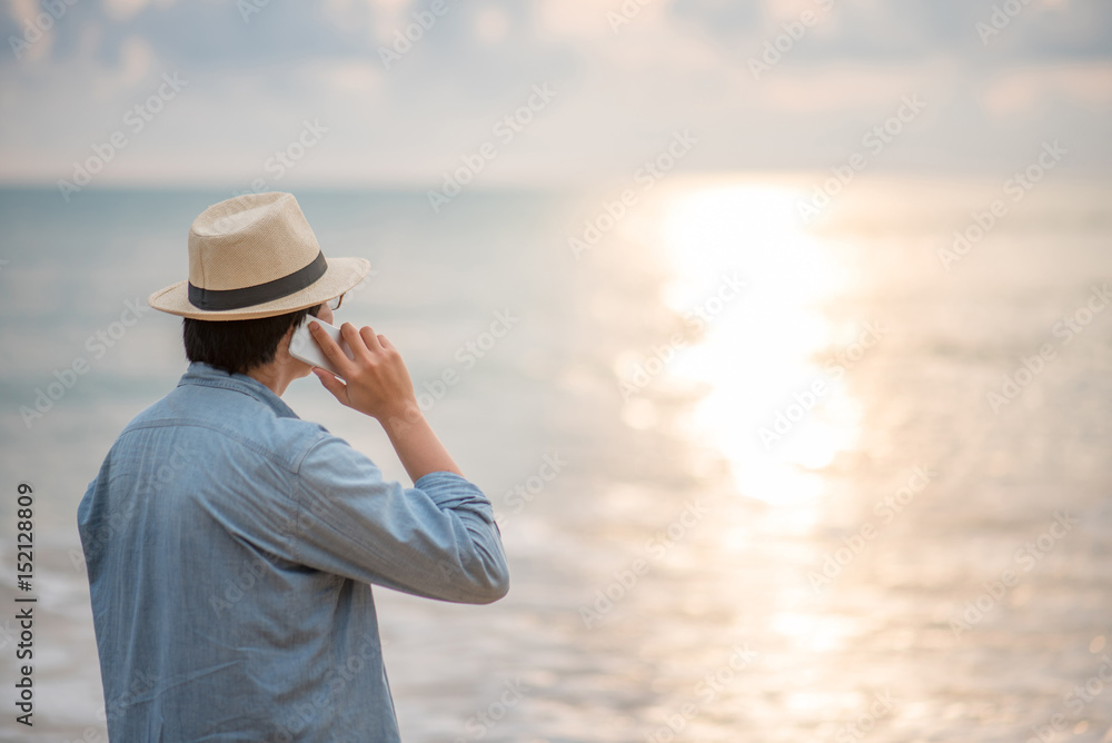 Young Asian man with jean shirt and hat talking on phone and see the sunset on tropical beach, background for summer holiday vacation or global communication concepts
