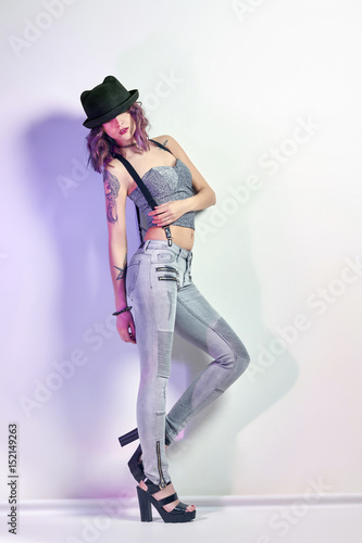 Sexy girl with purple hair and a tattoo on the body posing on grey background. Perfect woman in gray jeans and a t-shirt, bright beautiful makeup