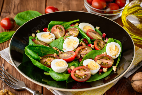 Fresh vegetable salad with spinach, cherry tomatoes, quail eggs, pomegranate seeds and walnuts in black plate on wooden table.