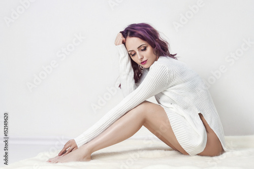 Sexy girl with purple hair and tattoos on his body posing in white shirt on gray background. Perfect woman in white dress sitting on the floor, bright beautiful makeup