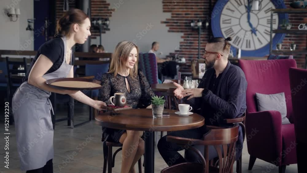 Hospitable young waitress bringing beverage in brown ceramic mug to the table for woman. Couple sitting at the restaurant together and talking. Colleagues having coffee break in cafeteria. Hospitable young waitress bringing beverage in brown ceramic mug to the table for woman. Couple sitting at the restaurant together and talking. Colleagues having coffee break in cafeteria.