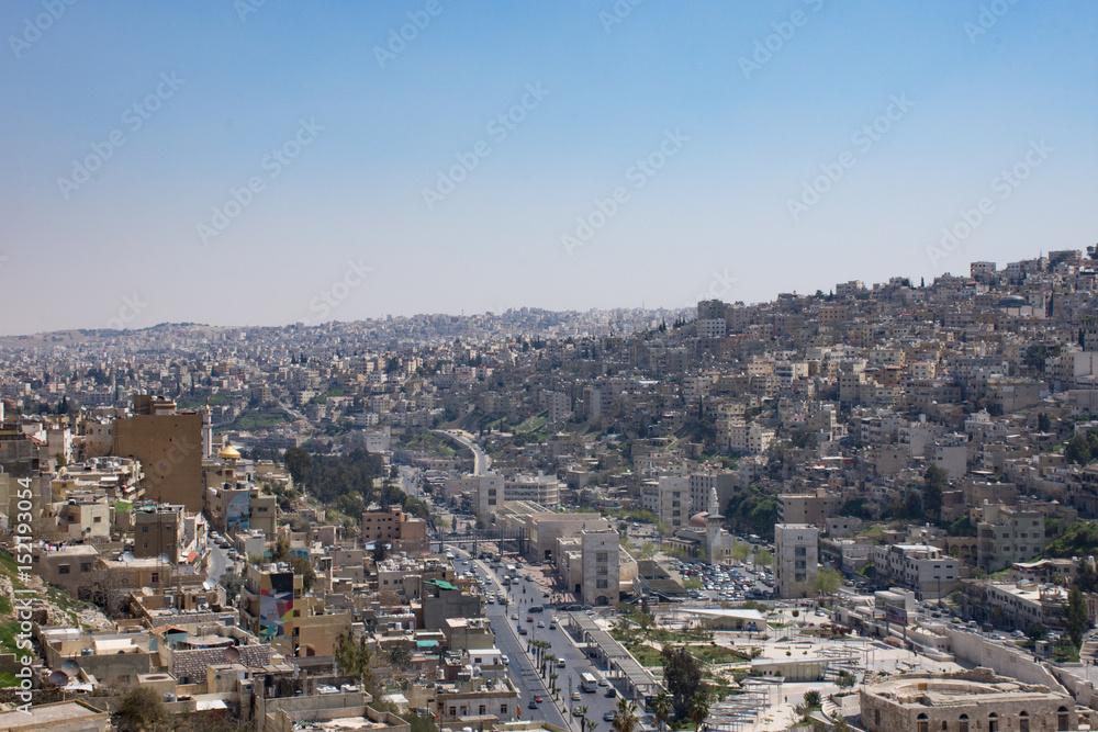 View of Amman's modern and older buildings including the Roman Theater below from the hill of Amman Citadel. Hazy blue sky is above.