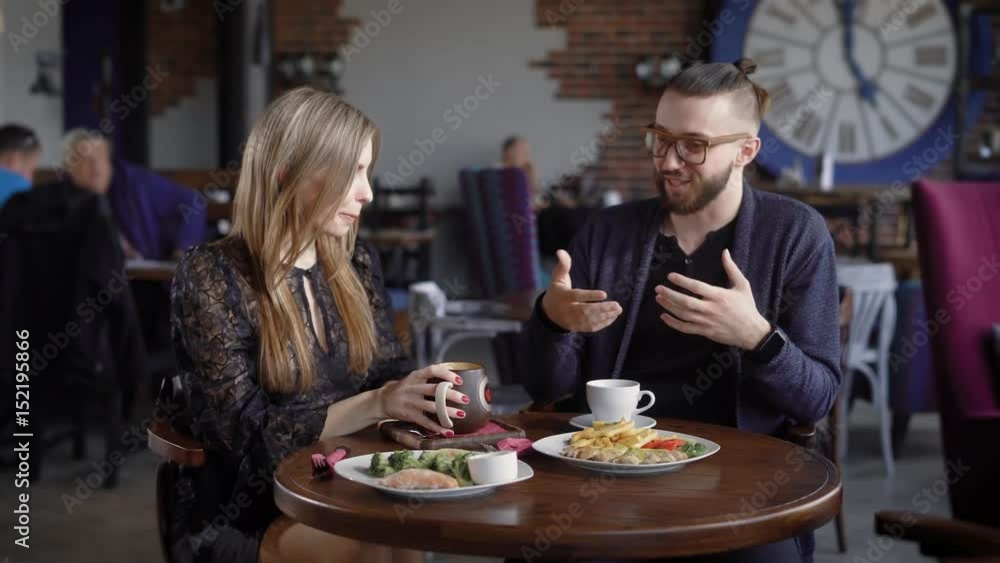 Happy couple of man and woman sitting in cafeteria at the served table with food and beverages spending time together. Beautiful lady drinking coffee listening to her gesticulating friend. Happy couple of man and woman sitting in cafeteria at the served table with food and beverages spending time together. Beautiful lady drinking coffee listening to her gesticulating friend.
