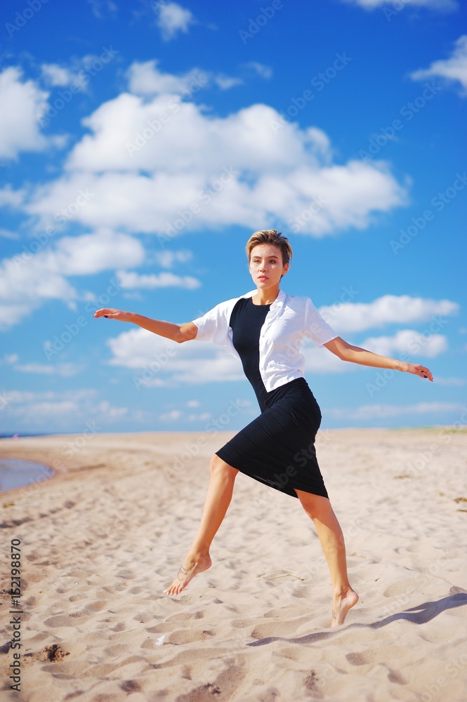 Portrait of a sad slender young woman with short hair, with outstretched hands, Bouncing on the sand, on blurred blue sky background Portrait of a sad slender young woman with short hair, with outstretched hands, Bouncing on the sand, on blurred blue sky background