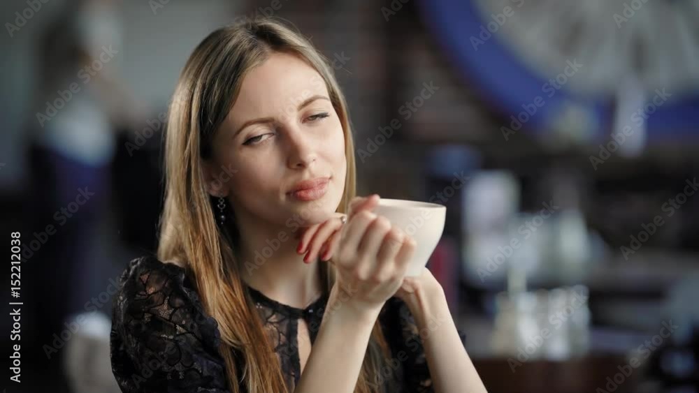 Beautiful woman sitting at the table in the restaurant with blurred background and holding cup of coffee. Adorable lady with natural make-up in black lace dress looking at the camera and smiling. Beautiful woman sitting at the table in the restaurant with blurred background and holding cup of coffee. Adorable lady with natural make-up in black lace dress looking at the camera and smiling.