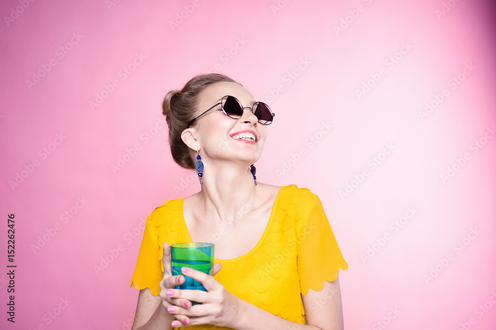 close-up portrait of a beautiful young blonde girl in fashionable sunglasses on a pink background in the studio in a yellow blouse with a blue glass cup of water in his hands with nail polish smiling