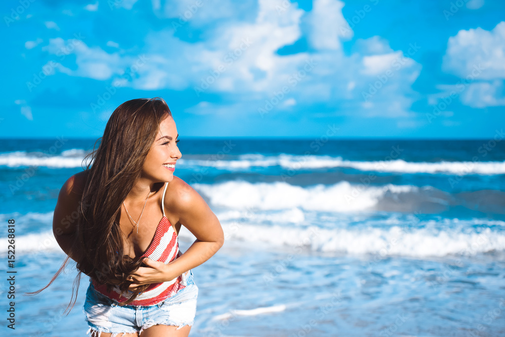 close-up portrait of a beautiful young brunette girl with long hair on a background of blue sea with waves and sky with clouds on a sunny day, lifestyle, posing and smiling, wind