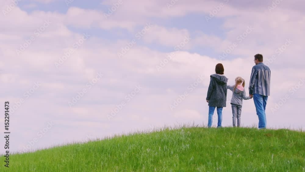 A family of three people - father, mother and daughter 6 years old, stand on top of a green hill. Looking forward to the horizon, rear view. Copyspace copmosition A family of three people - father, mother and daughter 6 years old, stand on top of a green hill. Looking forward to the horizon, rear view. Copyspace copmosition