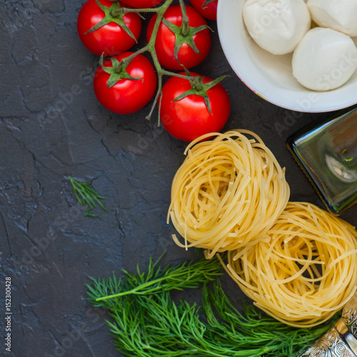 Ingredients for italian dinner. Olive oil, dill, cherry tomatoes, spices, mozzarella and italian whole grain pasta on a black slate background. Top view and copy space. Square image.