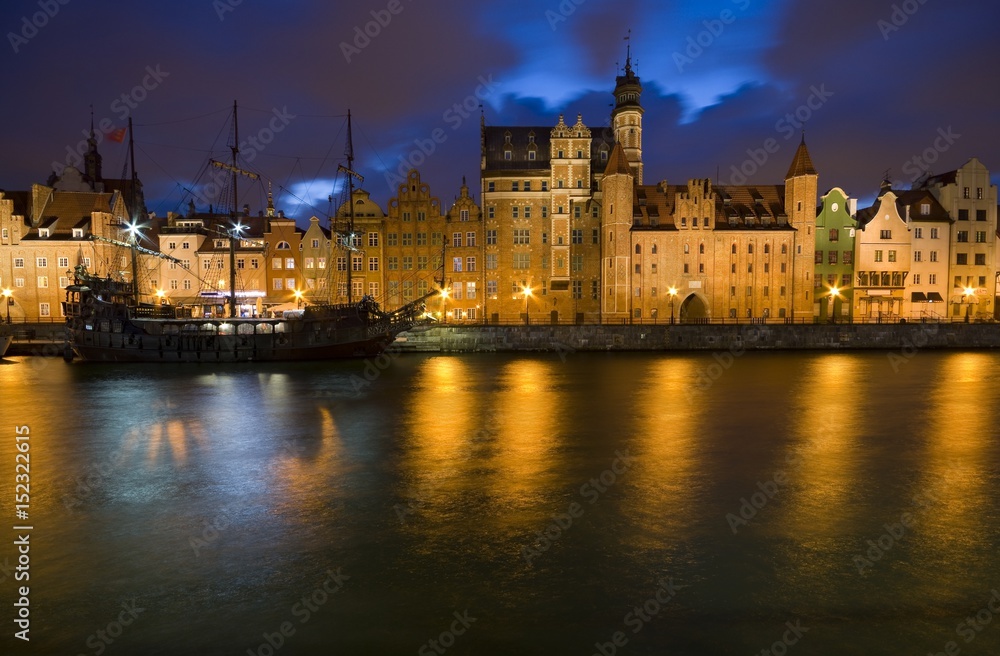 Caravel moored by Long Embankment on Motlawa River in the Old Town of Gdansk, Poland at night. Illuminated Mariacka Gate on the right