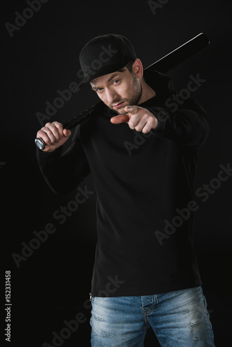 young robber in zoodie and cap with baseball bat isolated on black photo