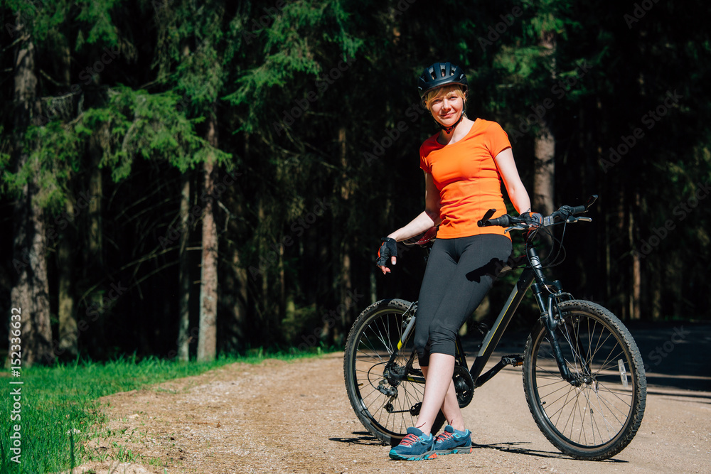 Obraz premium Young pretty woman in helmet and orange shirt with bicycle resting on the forest road after ride. Healthy lifestyle concept. Sports Girl with bike.