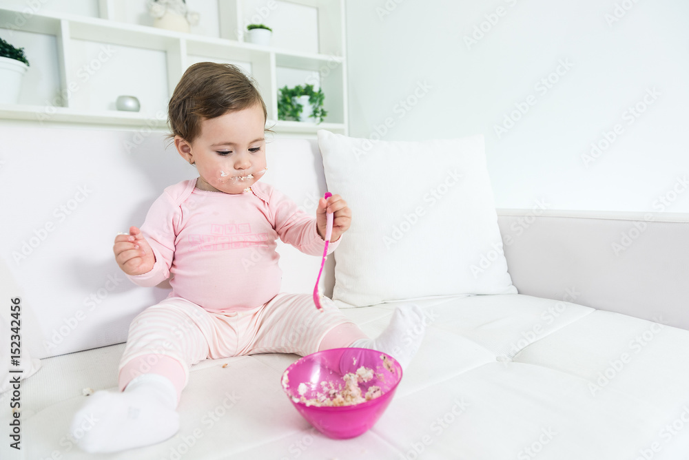 Happy baby little girl eating her first cake alone on the sofa.Shallow doff, copy space