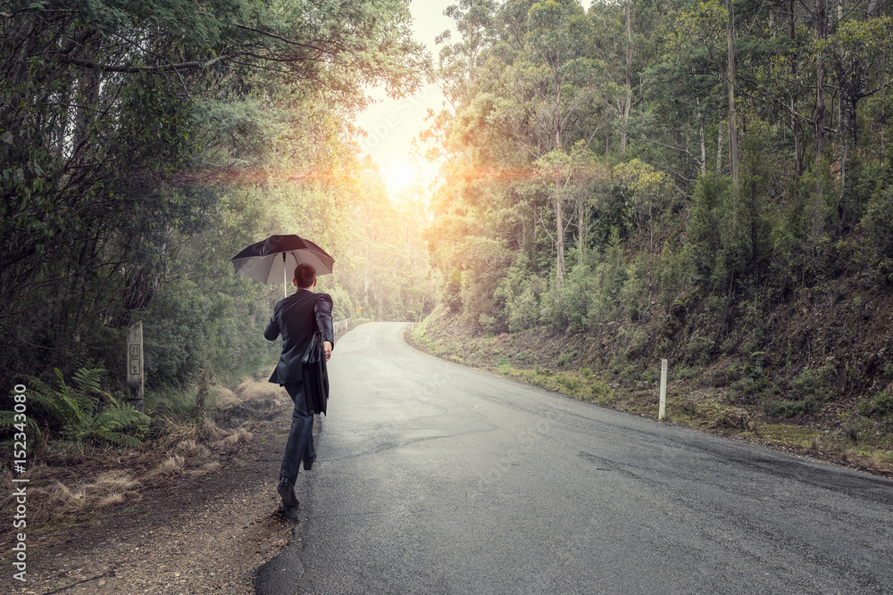 Obraz premium Back view of businessman with umbrella and suitcase walking on road