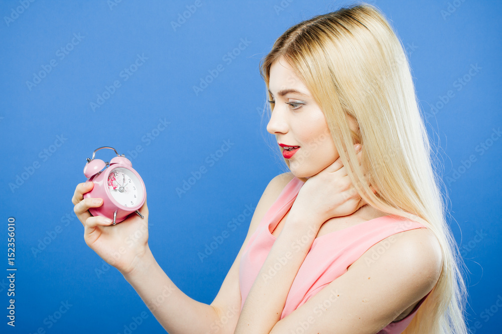 Surprised Young Blonde with Long Hair in Dress is Holding Pink Alarm Clock in Hand on Blue Background in Studio. Surprised Young Blonde with Long Hair in Dress is Holding Pink Alarm Clock in Hand on Blue Background in Studio.