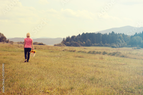 A young woman is walking across a large meadow on a sunny spring day and she is holding a wooden basket with sunflowers in her hand. © StockVideoFactory