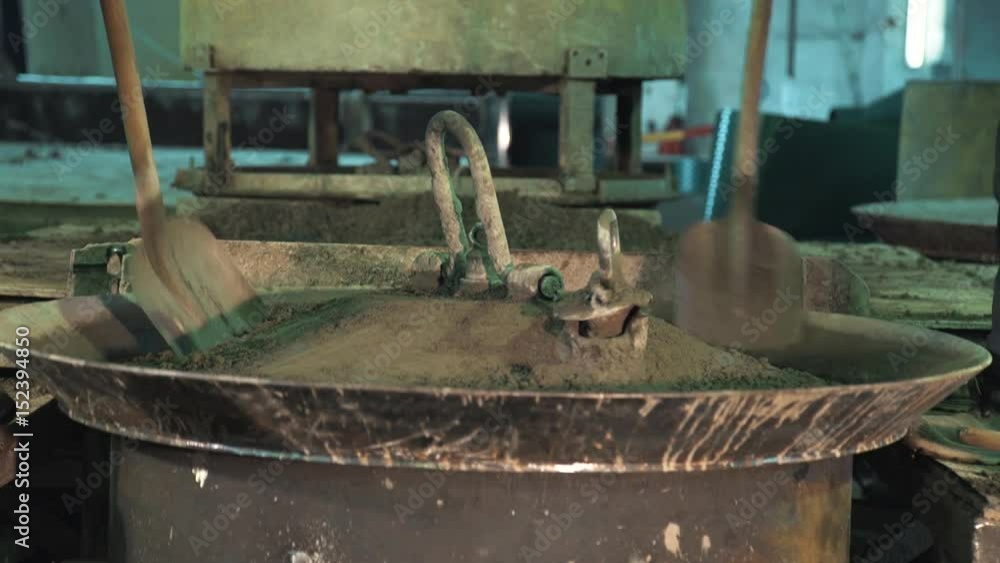 Two unrecognisable workers spread concrete with shovels over vibrating machine metal bowl at factory producing manholes Two unrecognisable workers spread concrete with shovels over vibrating machine metal bowl at factory producing manholes