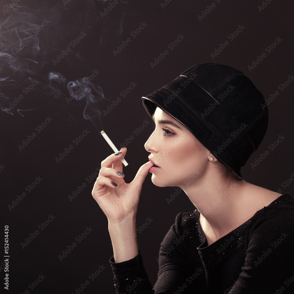 Obraz premium Young fashion woman smoking cigarette in hat and black drees over dark background. Vintage female portrait, styling. Image toned.