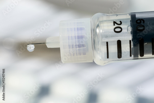 Close-up of pen's needle with a drop of insulin hanging on it. Inside the drop of the hormone are reflected other diabetes medicines and a notebook for blood tests results