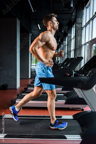 Side view full length of young man in sportswear running on treadmill at gym