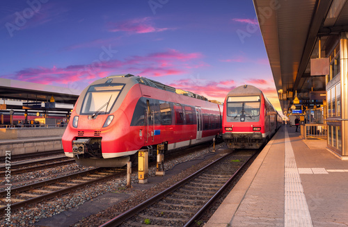 High speed red commuter trains on railway station and colorful sky at sunset in Europe. Industrial landscape with beautiful railway platform and modern trains in summer. Railroad. Railway tourism