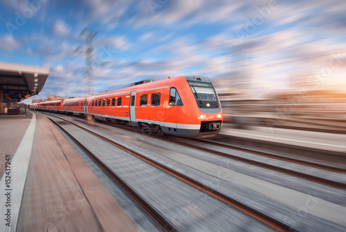 Red modern high speed train in motion on railroad track at sunset in Europe. Train on railway station with motion blur effect. Industrial landscape with train, railway platform and colorful sky