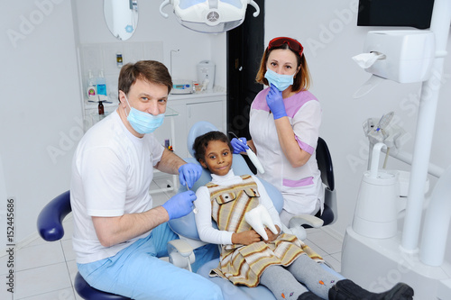 a dark-skinned baby girl in the dentist s chair. Two pediatric dentists and child in the dental office looking at the camera