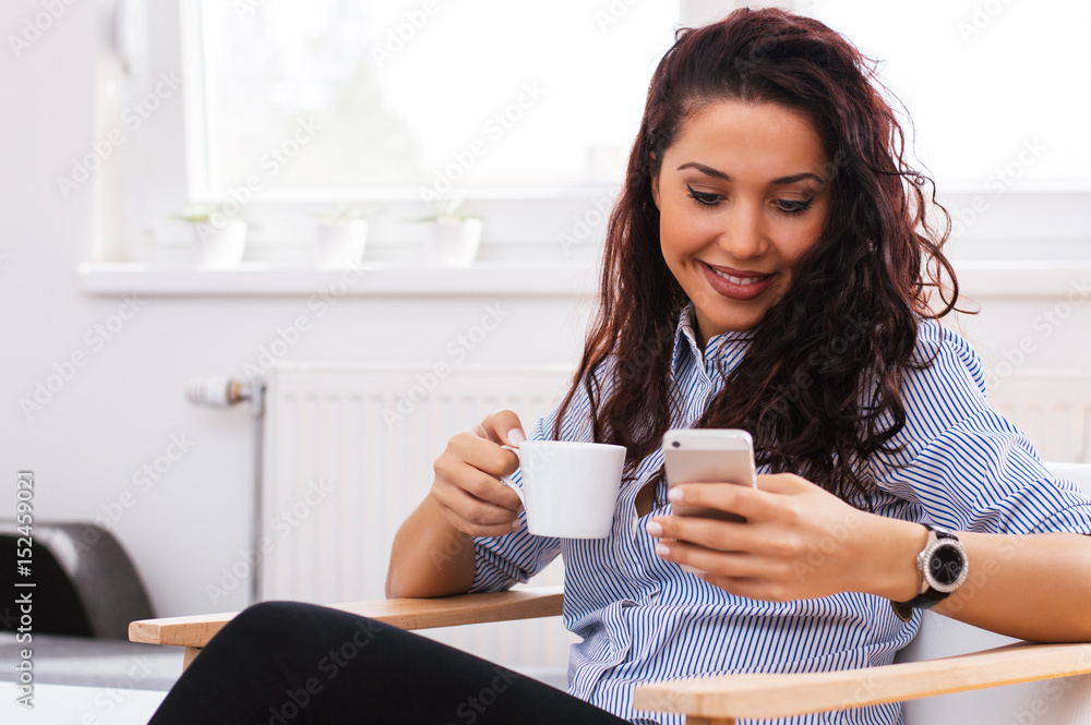 Portrait of young beautiful casual woman holding smartphone, looking at screen and holding cup of coffee Portrait of young beautiful casual woman holding smartphone, looking at screen and holding cup of coffee