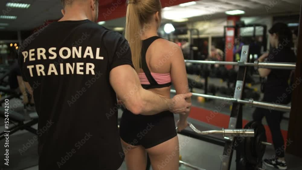 Personal trainer helping fit young female to lift curl bar, giving instructions Personal trainer helping fit young female to lift curl bar, giving instructions