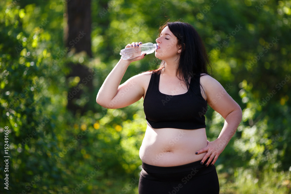 Young thirsty overweight woman drinking water after jogging in the park Young thirsty overweight woman drinking water after jogging in the park