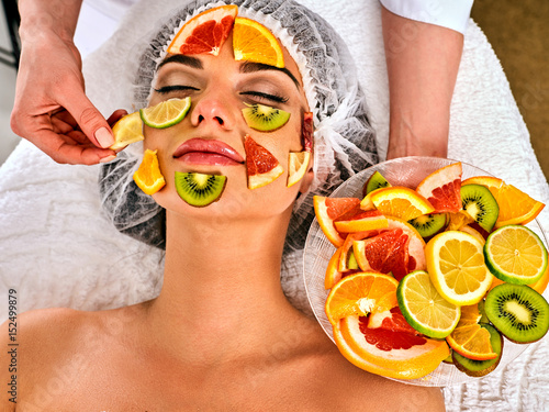 Facial mask from fresh fruits for woman . Beautician apply slices of avocado, grapefruit and kiwi Girl in medical hat lying on spa bed for skin care procedure in salon. Fruit salad for cosmetic mask.