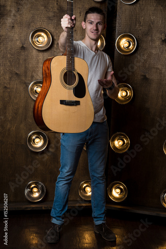 Guitarist, music. A young guy is standing with an acoustic guitar in his hand, in the background with lights behind him. Vertical frame