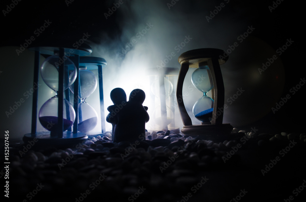 Time and love concept. Silhouettes of toy ceramic figures hugging between hourglasses in dark lighted background with fog Time and love concept. Silhouettes of toy ceramic figures hugging between hourglasses in dark lighted background with fog