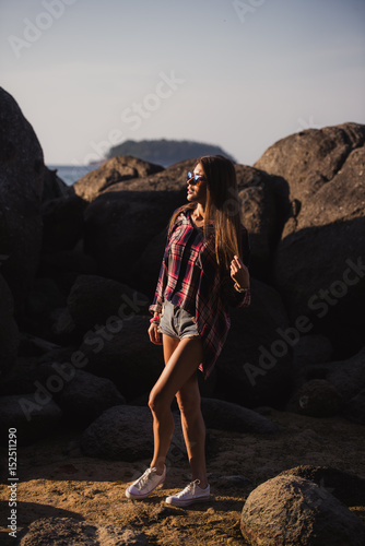 Beautiful slim woman in shirt, shorts and fashion sunglasses walk on big rocks. View from back. Beauty cute girl on a tropical beach sea ocean shore with large stones. Outdoor summer lifestyle.