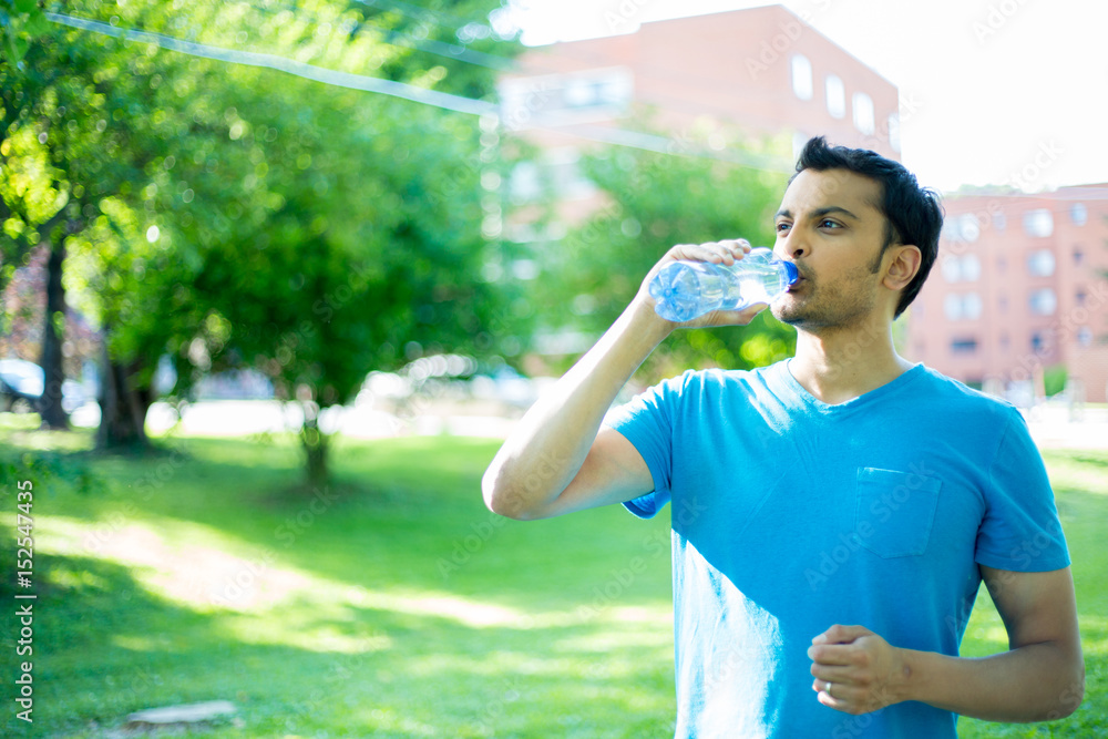 Closeup portrait of young guy in blue shirt drinking water from crystal clear bottle on a hot, sunny day, isolated green trees and building background Closeup portrait of young guy in blue shirt drinking water from crystal clear bottle on a hot, sunny day, isolated green trees and building background