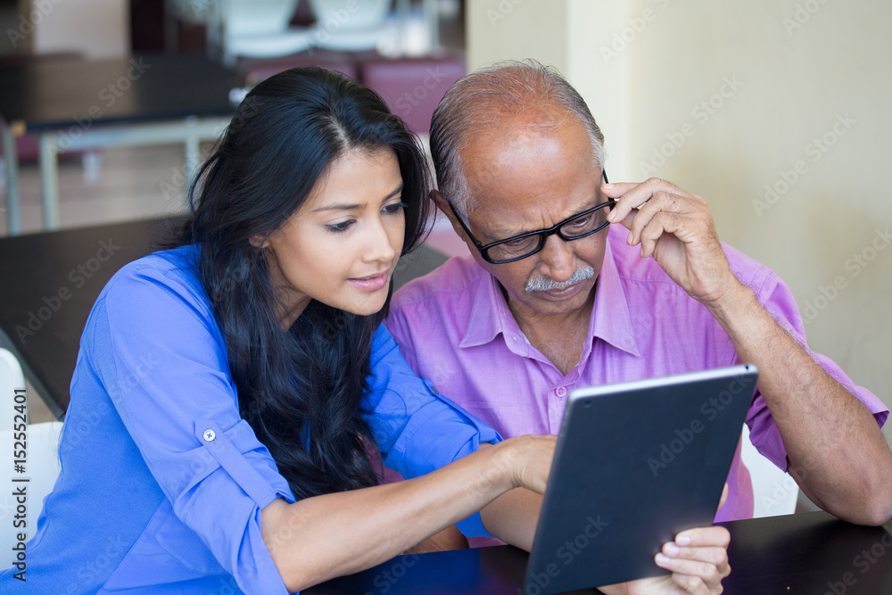 Closeup portrait, sitting young woman showing elderly with black glasses to use portable device,scrutinizing data with great concern, isolated indoors background Closeup portrait, sitting young woman showing elderly with black glasses to use portable device,scrutinizing data with great concern, isolated indoors background