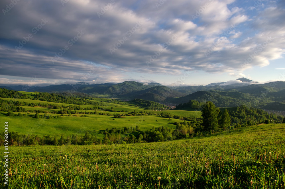 Valley among the green hills of the foothills with vivid bright meadows and groves of trees on the background of rural settlement and mountain ranges under the sunset sky.  Altai , Siberia, Russia.