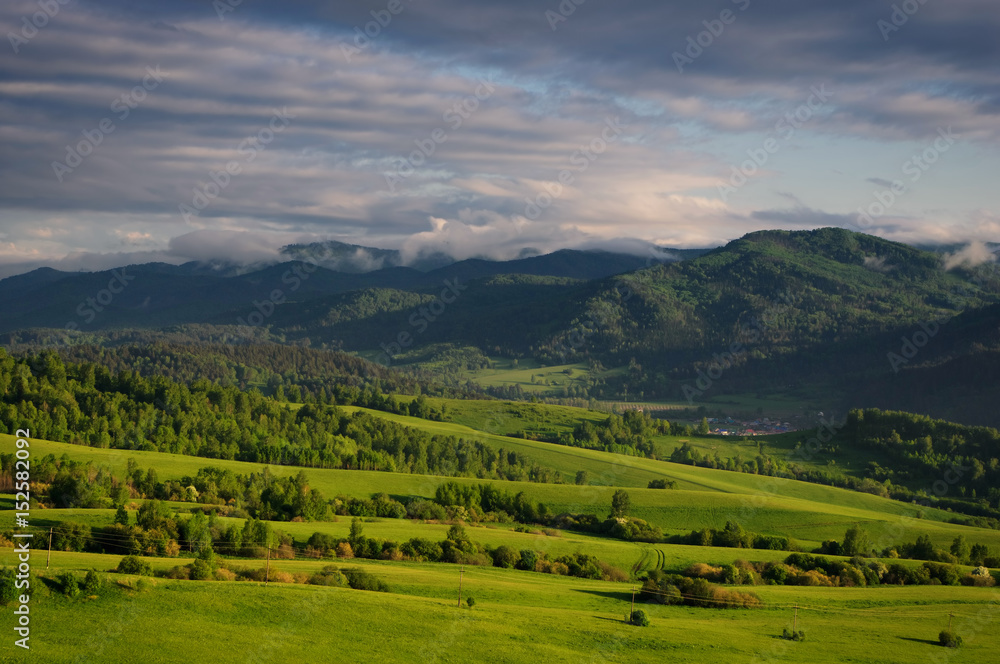 Valley among the green hills of the foothills with vivid meadows and groves of trees on the background of rural settlement and mountain ranges under the sunset sky.  Altai, Siberia, Russia.