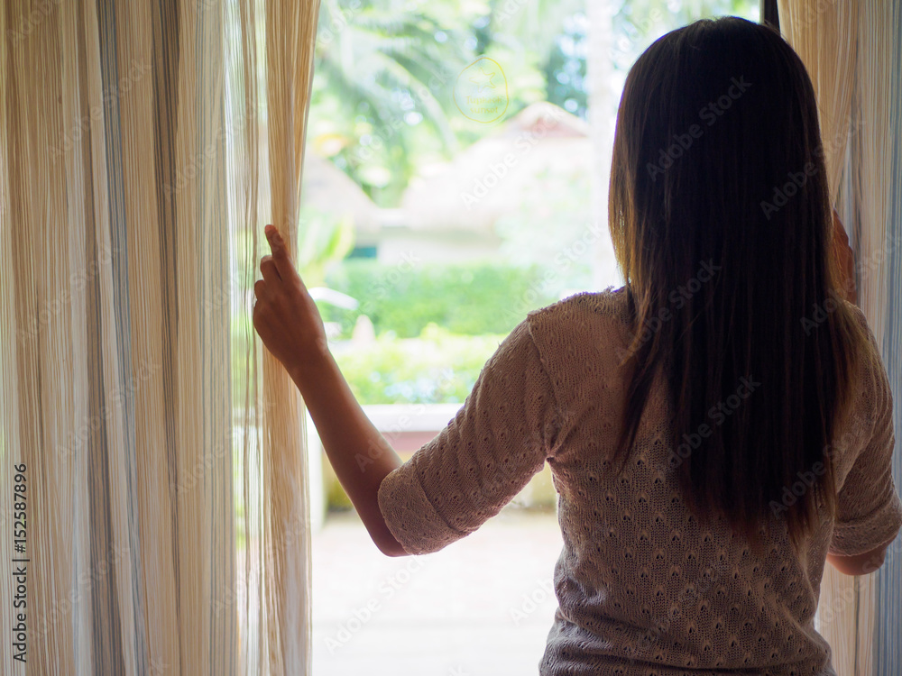 Rear view of a young woman holding the curtains open to look out of a large light window at home, interior. Positive and aspirational lifestyle. Sad Woman looking out a window, indoors.