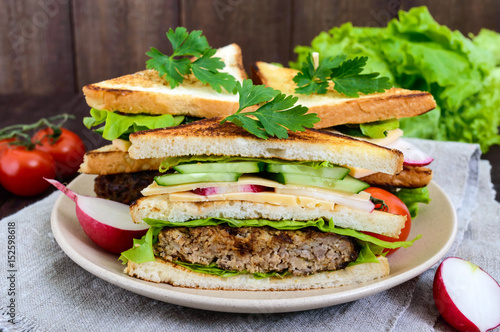 Multilayered sandwiches with a juicy cutlet, cheese, radish, cucumber, lettuce, arugula cutting in half on a plate on a dark wooden background.