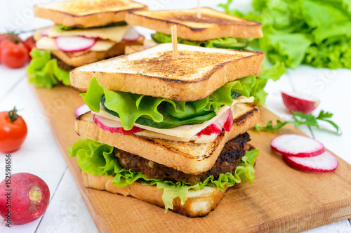 Multilayered sandwiches with a juicy cutlet, cheese, radish, cucumber, lettuce, arugula on a cutting board on a white wooden background.