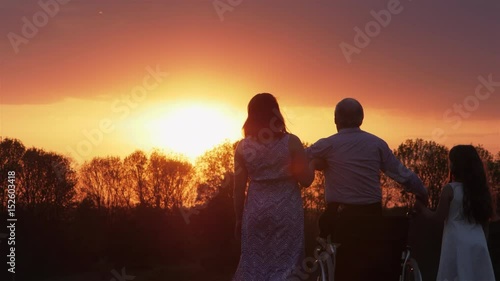 Disabled father with daughter and granddaughter watching the sunset. An elderly man gets up from a wheelchair