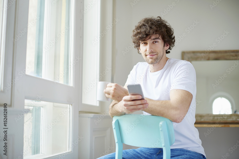 Starting the day with coffee and reading messages. Shot of a young man having a cup of coffee and text messaging while looking out his apartment window.
