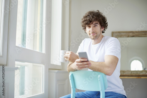Starting the day with coffee and reading messages. Shot of a young man having a cup of coffee and text messaging while looking out his apartment window.