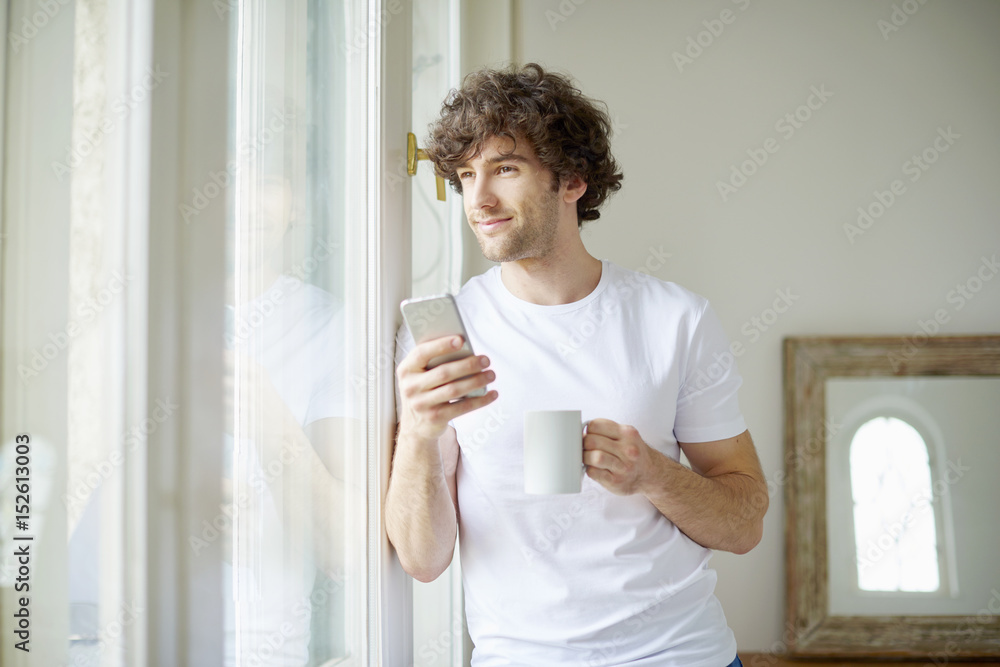 Starting the day with coffee and reading messages. Shot of a young man having a cup of coffee and text messaging while looking out his apartment window. Starting the day with coffee and reading messages. Shot of a young man having a cup of coffee and text messaging while looking out his apartment window.
