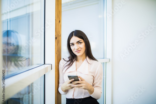 Young woman busy using her mobile phone while standing in front of large glass windows