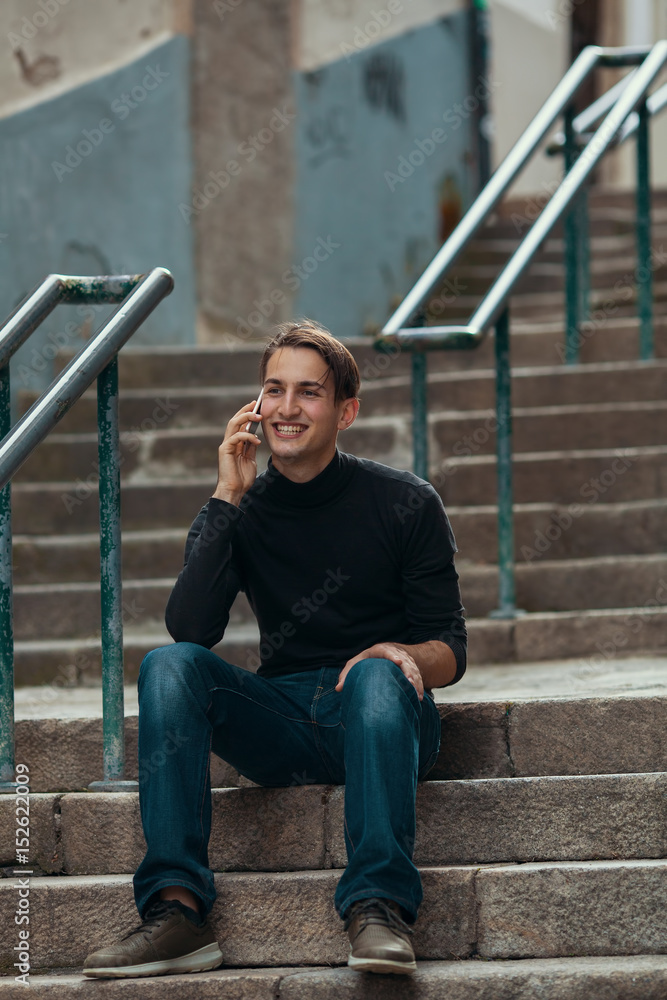 Handsome young man talking on mobile phone while sitting on stone steps outdoors.