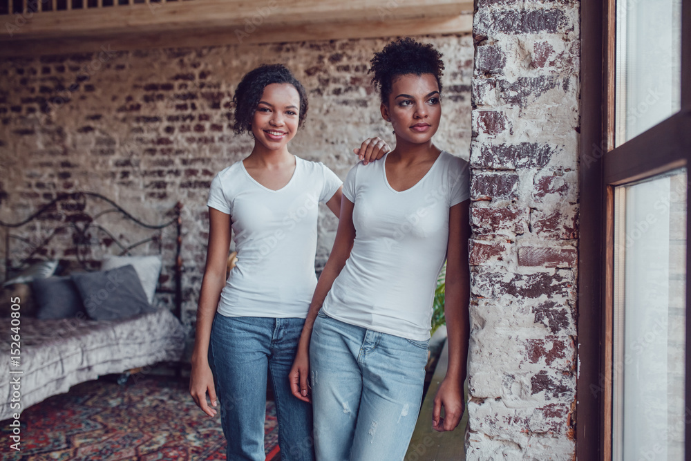 Two attractive african american girls in white T-shirts are standing near the window. Two attractive african american girls in white T-shirts are standing near the window.