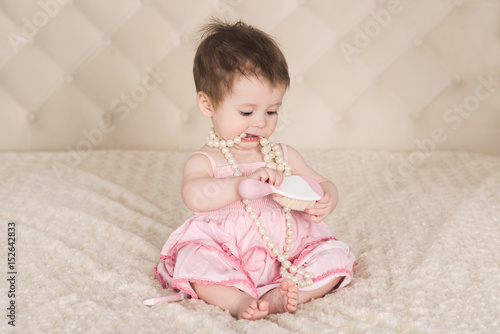 cute baby girl in pink dress, with pearl beads and comb in her hands sitting on the bed and playing, indoors